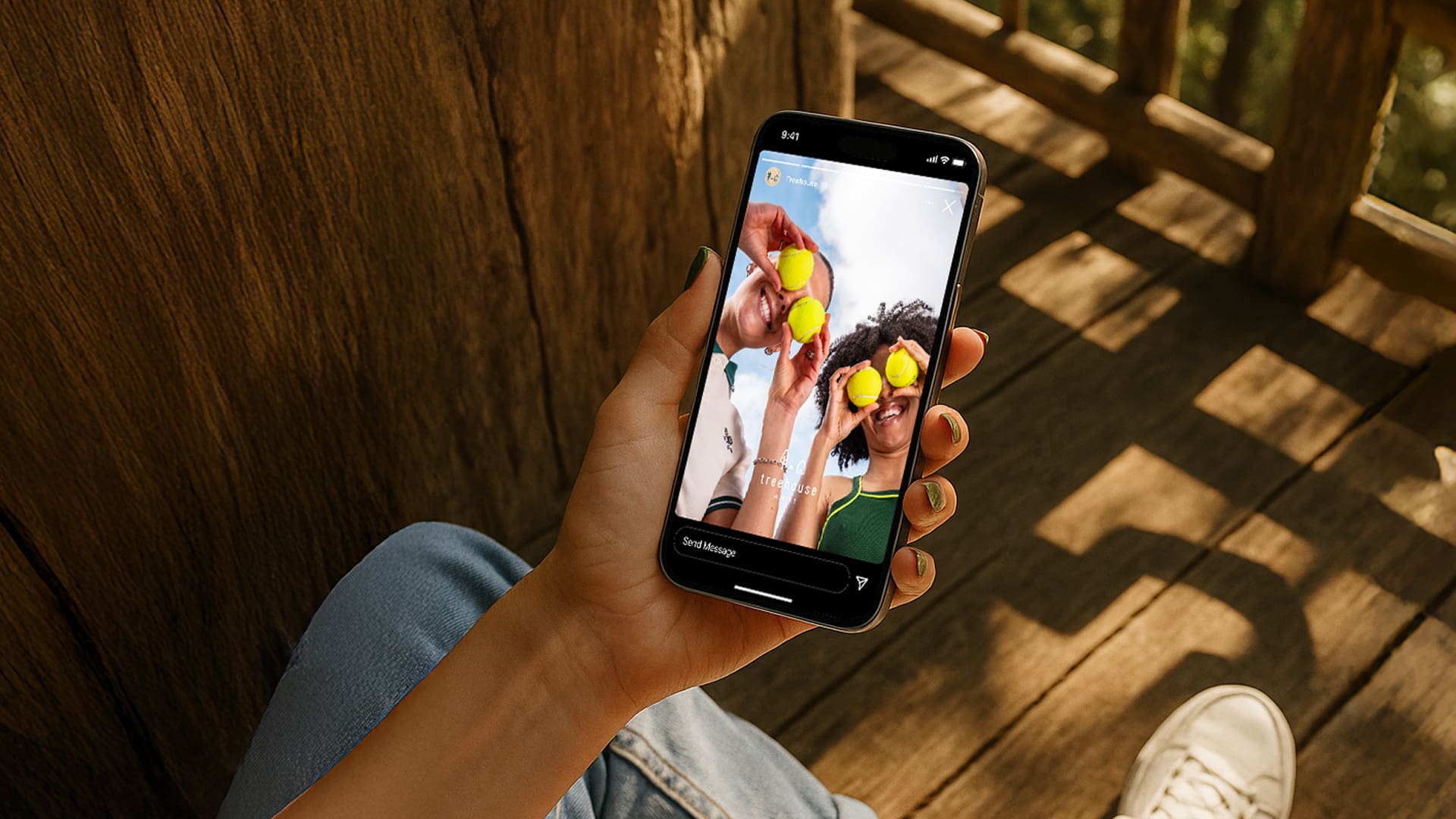 A person sits outdoors on wooden steps, holding a smartphone showing a photo of two people playfully holding lemons over their eyes and smiling. Sunlight casts warm shadows on the scene.
