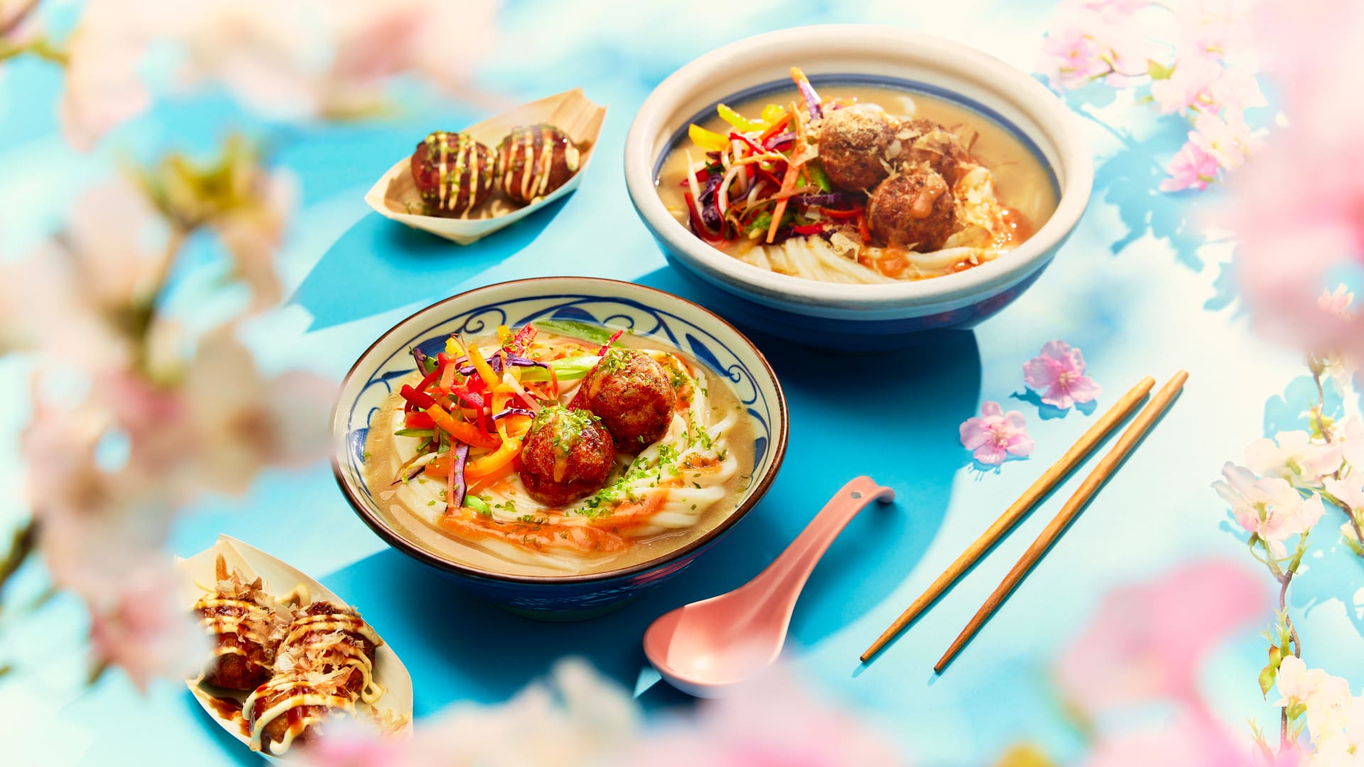 Two colorful bowls of noodle soup topped with vegetables and meatballs are set on a blue table with chopsticks and a pink spoon. Two small plates of takoyaki are on the side, surrounded by pink cherry blossom flowers.