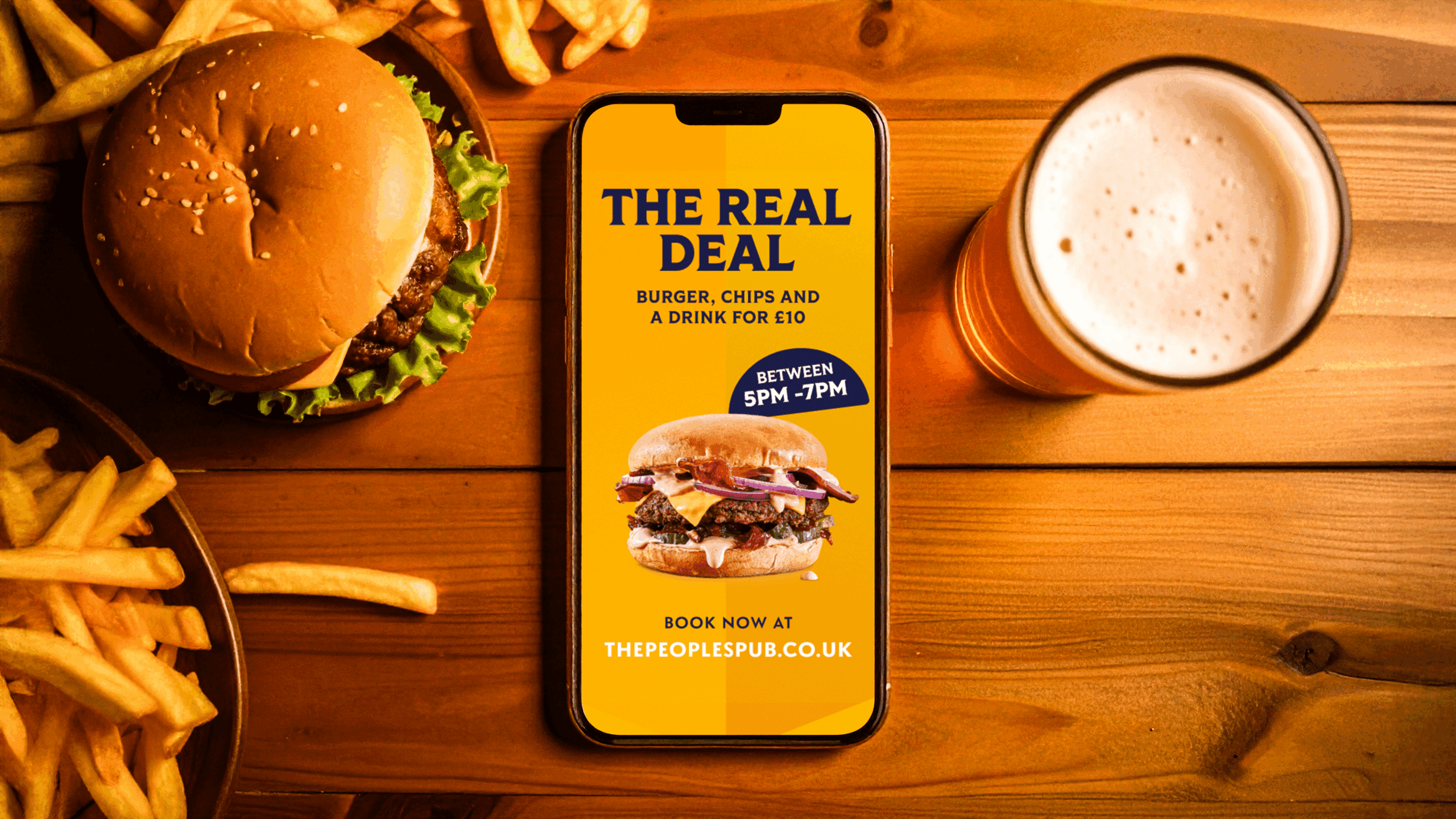 A burger, fries, and a pint of beer on a wooden table. A phone displays a yellow ad offering a burger, chips, and a drink for £10 at The Peoples Pub, available 5-7pm.