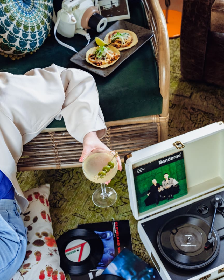 A person relaxes on a pillow near a wicker sofa, holding a martini with olives. Two tostadas are on a plate, and a record player spins a vinyl next to scattered records on the patterned green carpet.