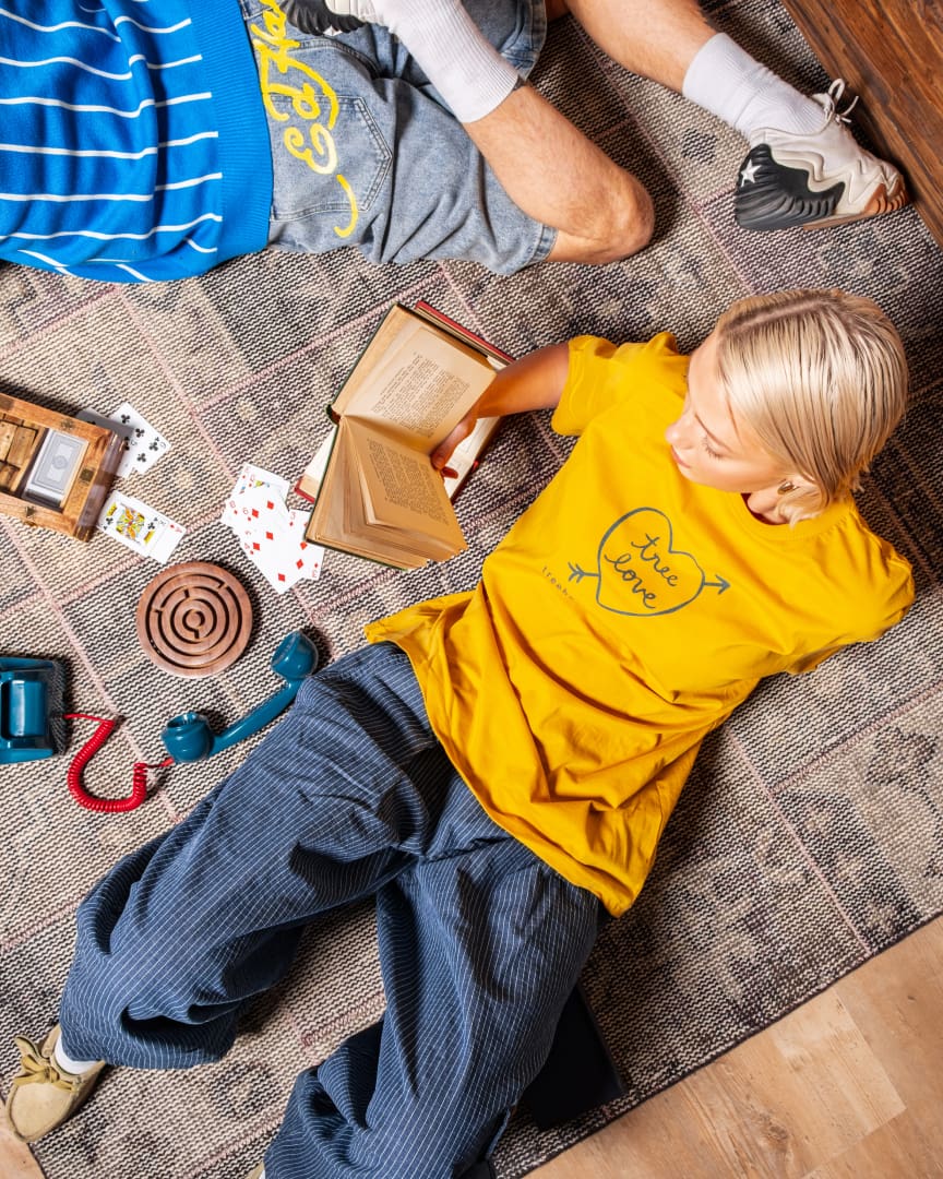 Two people lie on a rug surrounded by playing cards, an open book, a rotary phone, headphones, and a tape recorder. One person wears a yellow "True Love" shirt and blue pants; the other wears a striped shirt and gray shorts.