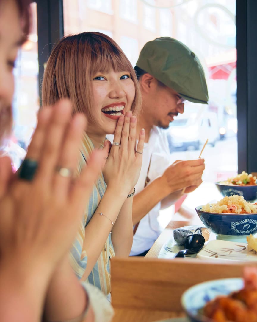 A woman smiles and clasps her hands joyfully at a restaurant table, with bowls of food in front of her. A man beside her, wearing a green cap, is focused on his meal. The setting appears casual and lively.
