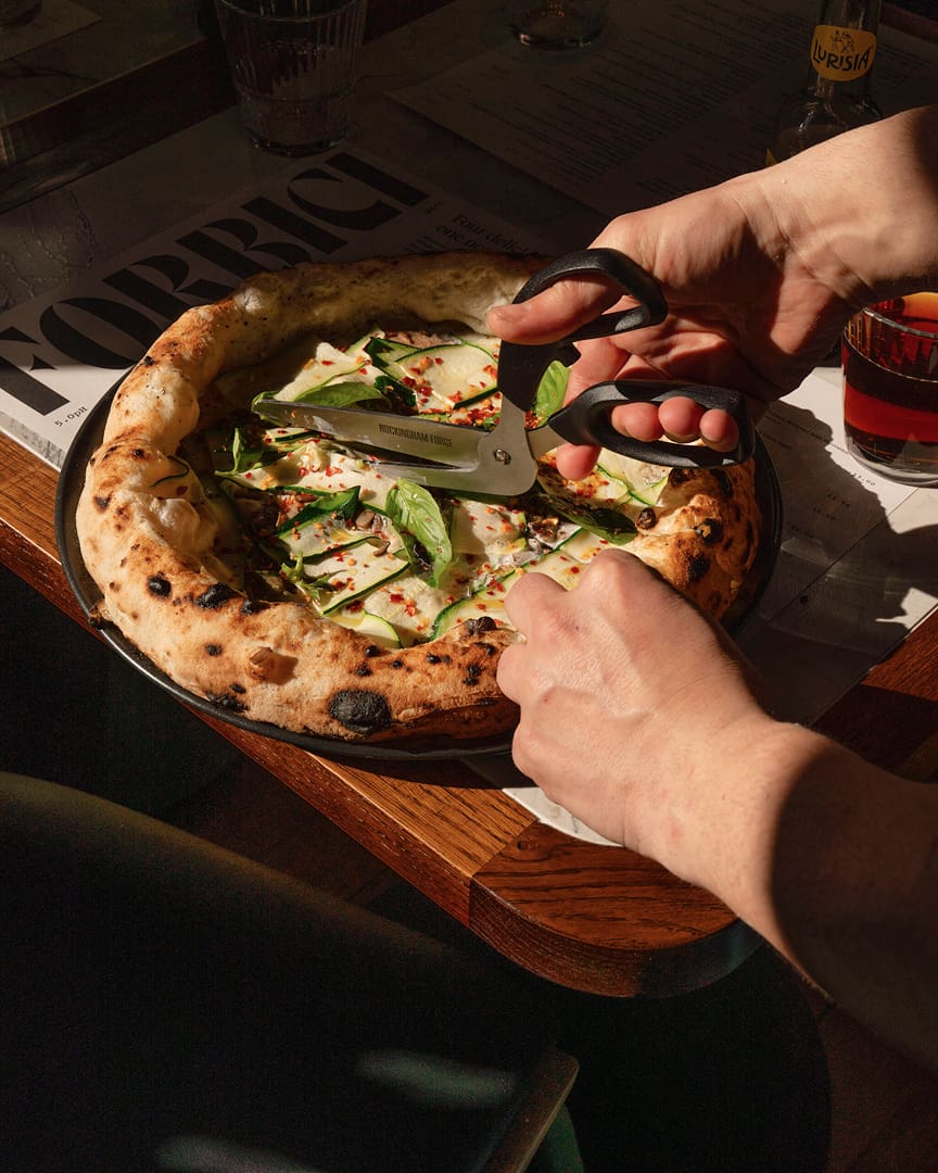A person uses scissors to cut a pizza topped with fresh basil and red pepper flakes, with sunlight casting shadows on the pizza and hands. A newspaper, drink, and glass are visible on the wooden table.