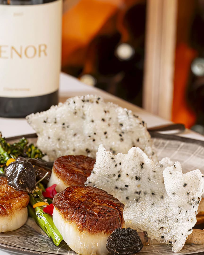A plate with seared scallops, grilled vegetables, crispy sesame crackers, and dark mushrooms, with a bottle of white wine in the background.