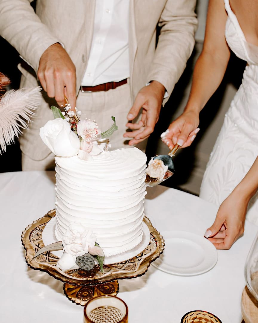 A bride and groom in elegant attire cut a white, textured wedding cake decorated with flowers, placed on a decorative stand, as they prepare to serve a slice on a plate.