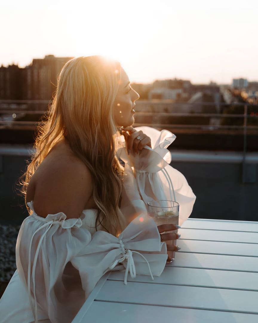 A woman with long, wavy hair in an off-the-shoulder white dress sits at a white table outdoors, holding a drink, with sunlight streaming over her at sunset and a cityscape in the background.
