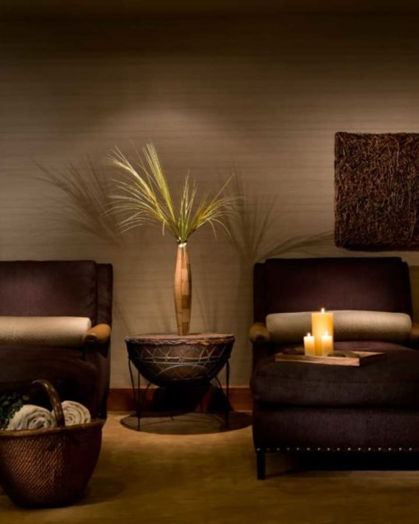 Two dark armchairs with beige cushions sit on either side of a table with a decorative vase and grass. Candles on a tray, a basket with rolled blankets, and dim, warm lighting create a cozy atmosphere.