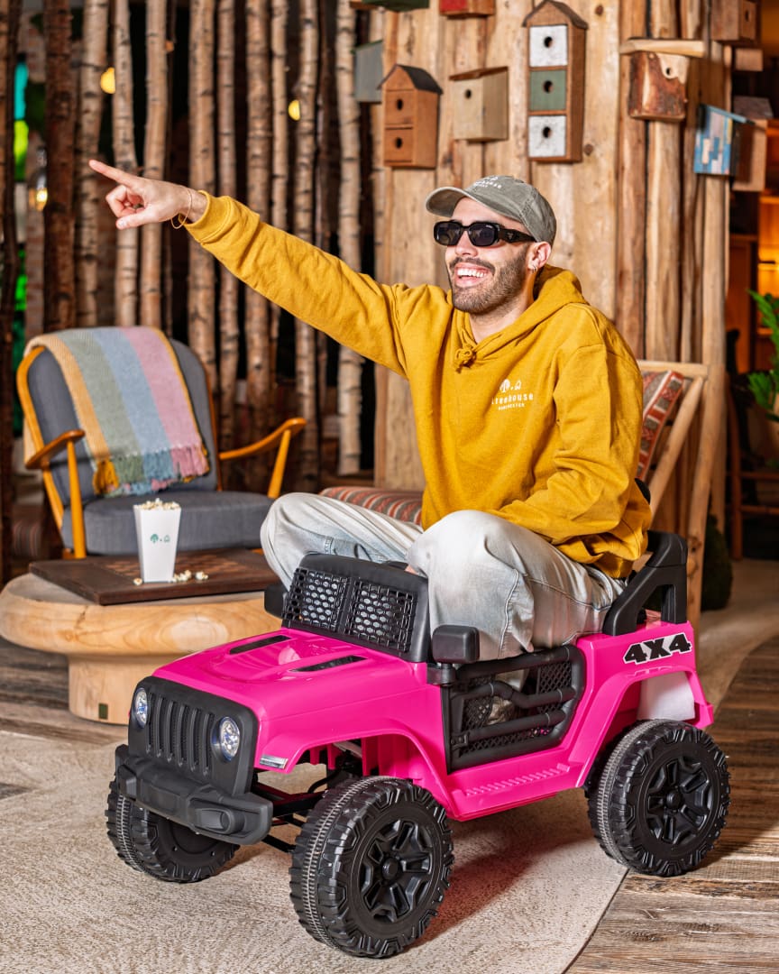 A smiling man in sunglasses and a yellow hoodie sits on a small pink toy 4x4 car indoors, pointing excitedly to the side. The cozy room has wooden decor, birdhouses, and a colorful striped blanket on a chair.