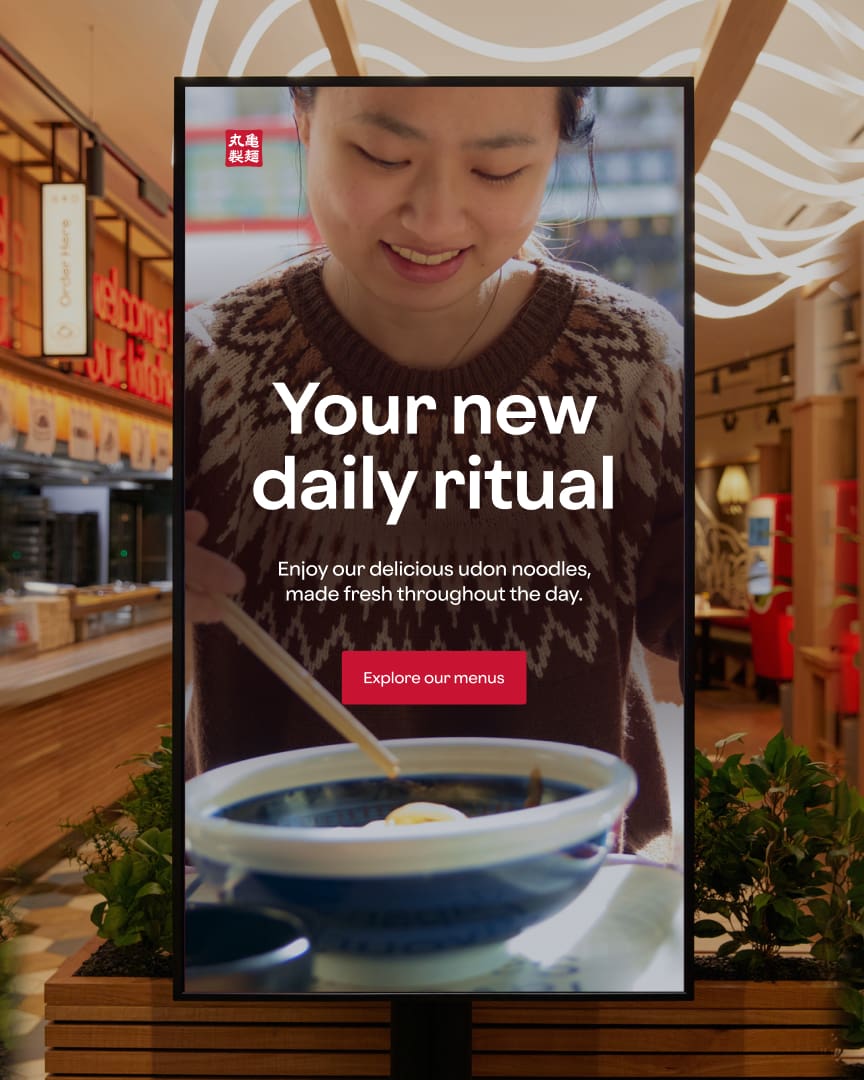 A restaurant sign shows a woman smiling at a bowl of udon noodles. Text reads: "Your new daily ritual. Enjoy our delicious udon noodles, made fresh throughout the day." A button below says, "Explore our menus.