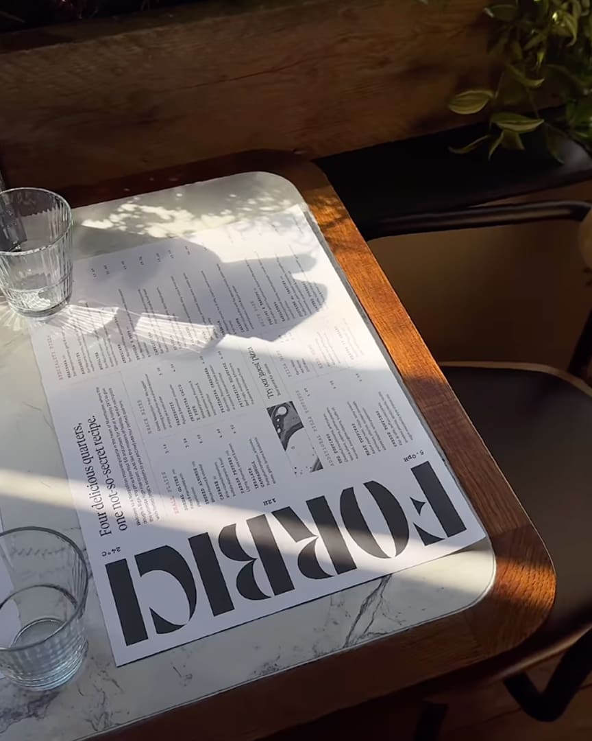 A sunlit table with a "FORBICI" menu, two empty glasses, and a plant nearby. Shadows from the glasses and plant fall across the marble tabletop and menu, creating a cozy café atmosphere.