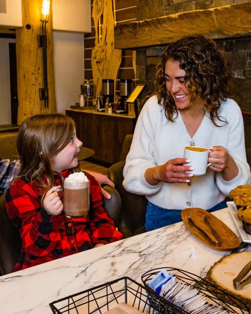 A woman and a young girl sit at a marble table, smiling at each other. The girl holds a mug of hot chocolate with whipped cream; the woman holds a cup of coffee. Pastries are on the table in a cozy, rustic setting.
