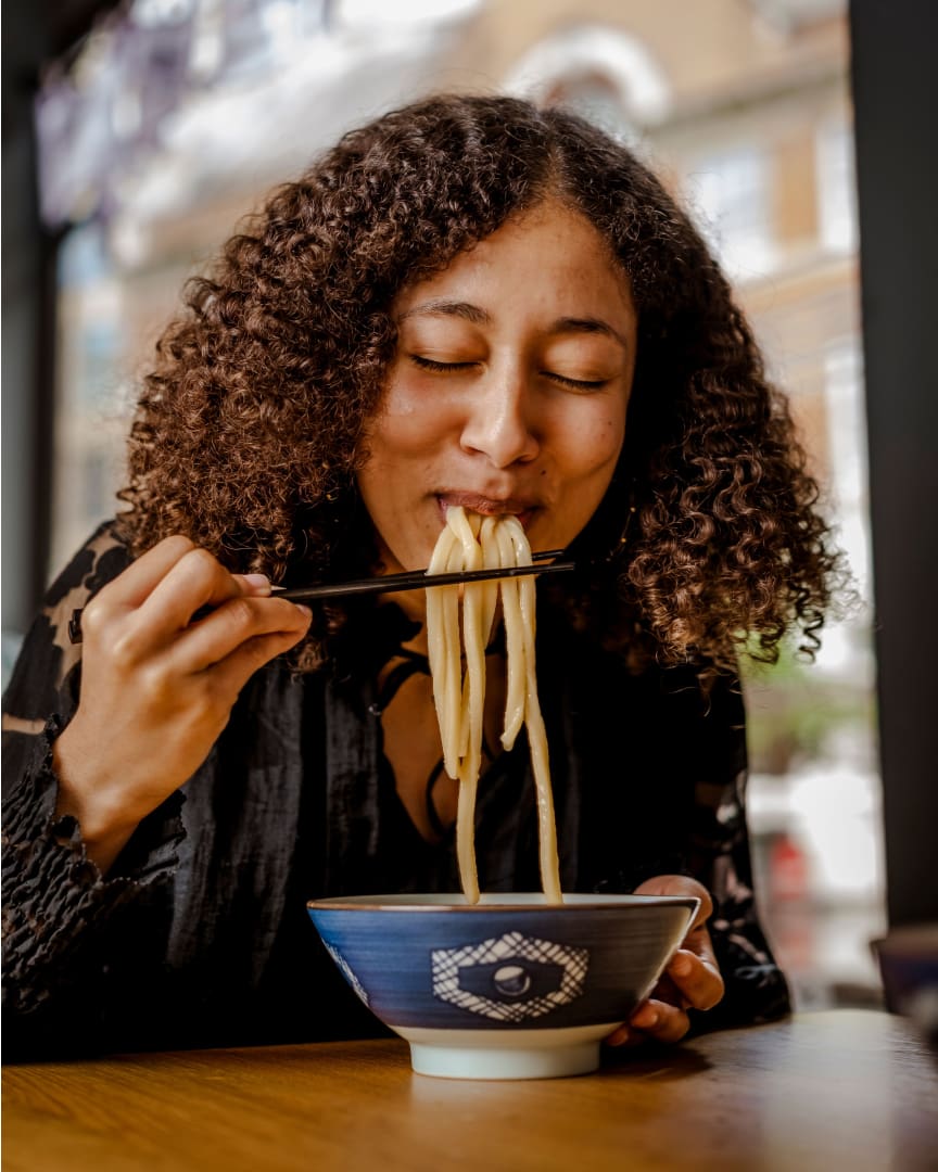 A woman with curly hair smiles while eating noodles with chopsticks from a bowl, sitting at a table in a restaurant with a blurred street scene visible through the window behind her.
