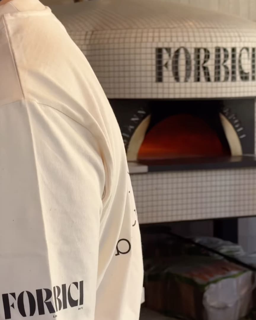 A person wearing a white shirt with "FORBICI" printed on the sleeve stands in front of a tiled pizza oven with "FORBICI" displayed on its front. The oven's interior glows with orange light.