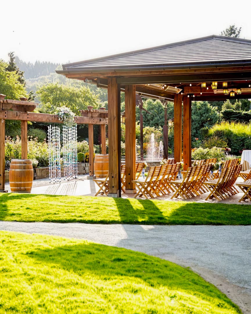 Rows of wooden chairs are arranged under a wooden pavilion in a sunny outdoor garden setting, with lush greenery, a barrel, and decorative features visible in the background.