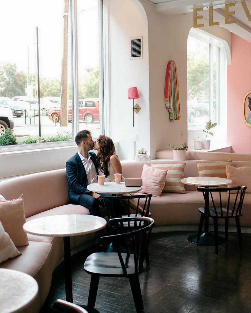 A couple sits closely together on a pink curved sofa in a bright, modern café, sharing a kiss at a small marble table. The room features pink and white decor, large windows, and minimalistic furnishings.