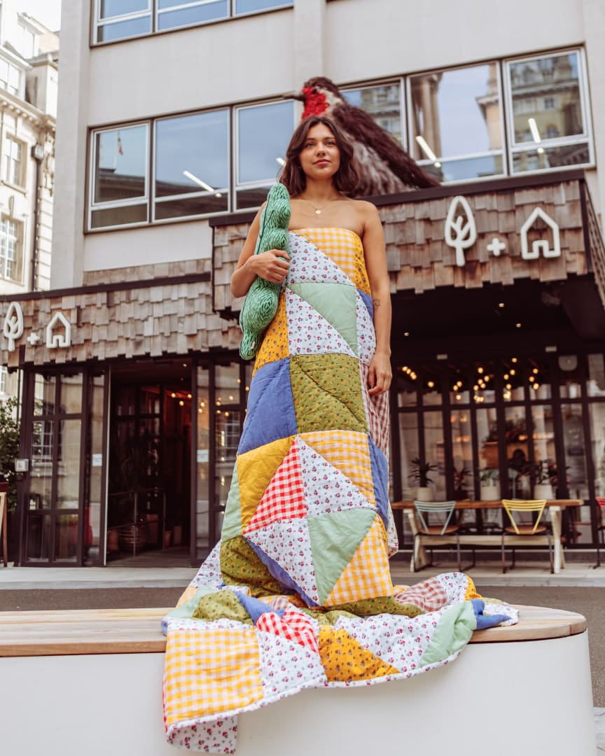 A woman stands outdoors wrapped in a colorful patchwork quilt, holding a green plush object. She is in front of a modern building with wood accents and large windows.