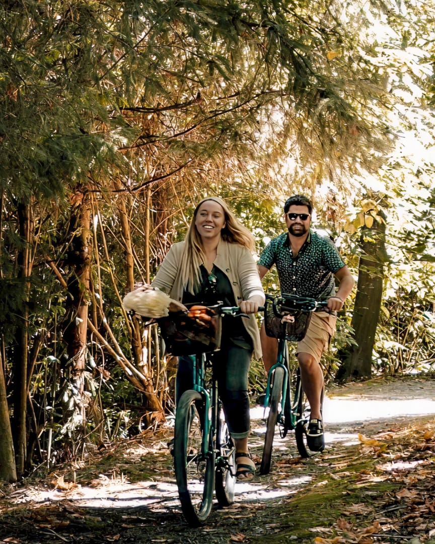 A smiling woman and man ride bicycles along a sunlit, wooded path. The woman leads with a basket on her bike, and both appear happy and relaxed, surrounded by lush green foliage.