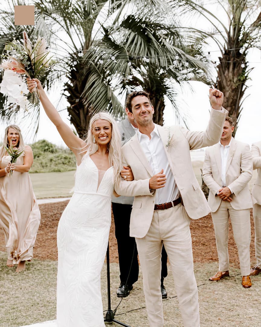 A bride and groom celebrate outdoors, smiling with arms raised, under palm trees. The bride holds a bouquet and wears a white dress; the groom wears a light suit. Bridesmaids and groomsmen in beige attire stand behind them.