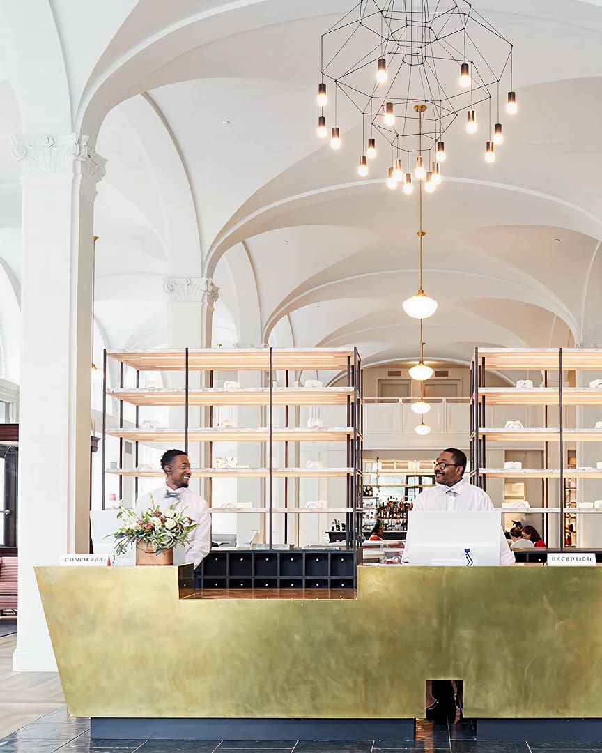 A modern hotel lobby with two men standing behind a gold reception desk. Behind them are shelves with neatly arranged items. The space features white vaulted ceilings and a geometric chandelier with hanging lights.