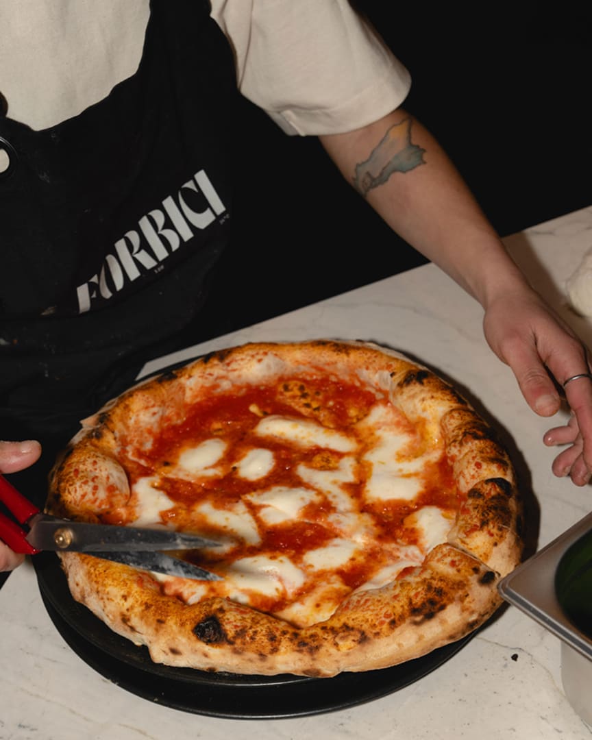 A person wearing a black apron with "FORBICI" on it slices a freshly baked Neapolitan pizza with melted cheese and tomato sauce on a white marble counter.