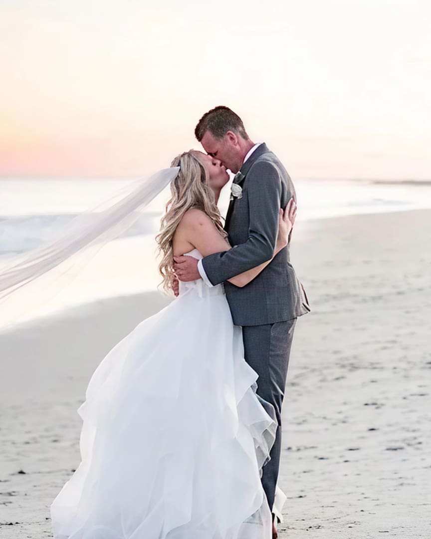 A bride and groom kiss on a sandy beach at sunset. The bride wears a long white gown with a veil flowing in the breeze, and the groom is dressed in a gray suit. The ocean and soft pastel sky are in the background.