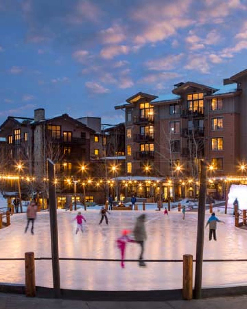 People ice skating on an outdoor rink at dusk, surrounded by string lights and modern apartment buildings with illuminated windows under a partly cloudy sky.