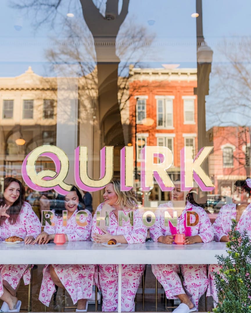 Five women in matching pink floral pajamas sit at a counter with coffee and pastries, smiling and talking, seen through a window with "QUIRK RICHMOND" in large letters. Reflections of buildings and trees appear in the glass.