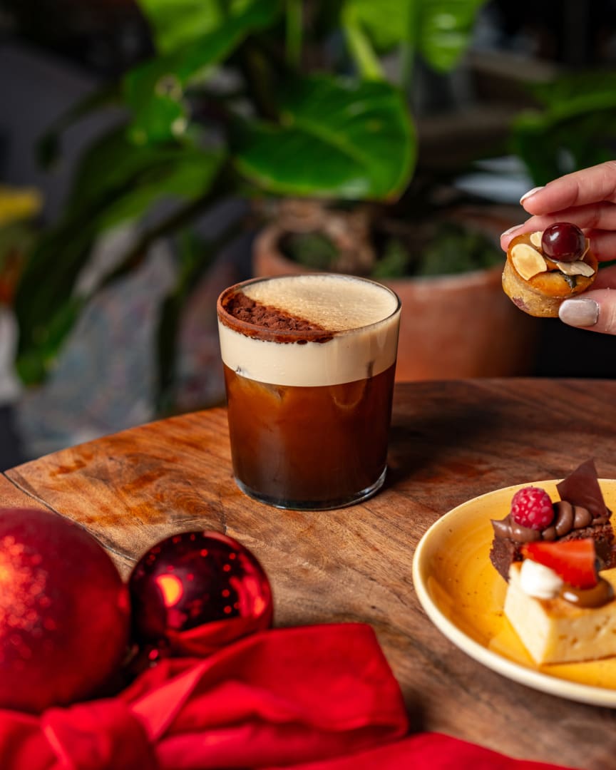 A glass of iced coffee with foam sits on a wooden table near holiday ornaments and desserts. A hand holds a small pastry over a yellow plate with assorted sweets. Green plant leaves are in the background.