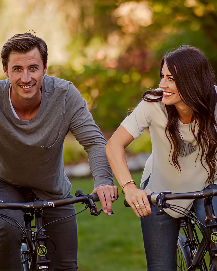 A man and a woman smiling while riding bicycles together outdoors on a grassy area with trees in the background. The woman looks at the man as they enjoy the activity.
