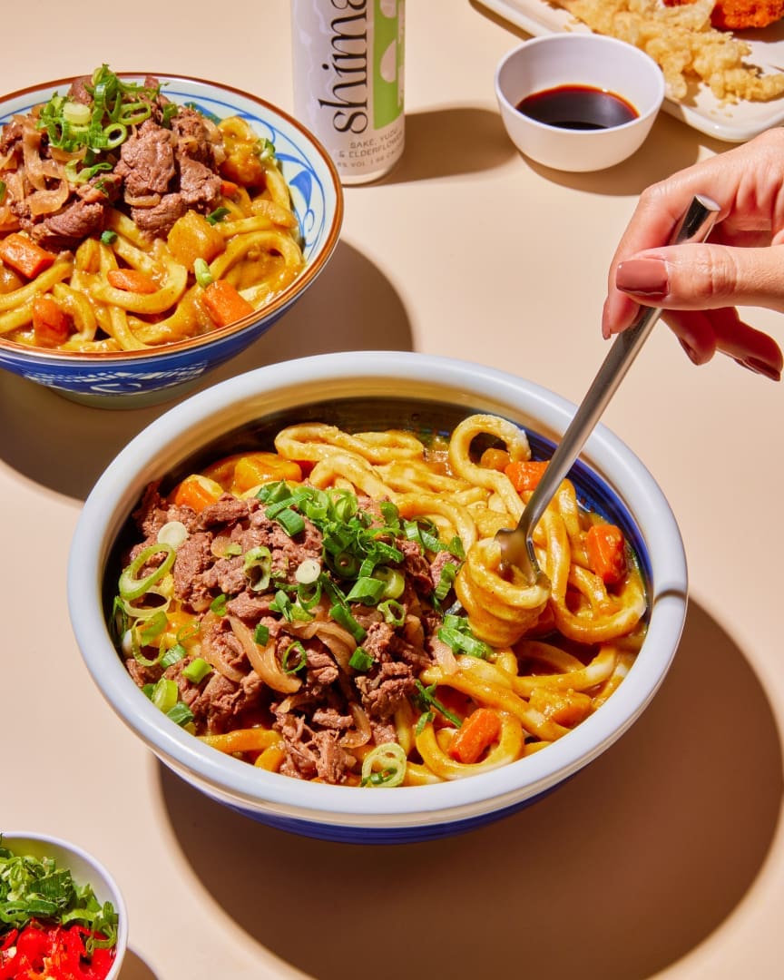 A hand uses chopsticks to lift thick udon noodles from a bowl topped with sliced beef, scallions, and vegetables. Another similar bowl, a drink can, and small dishes of sauce are also on the table.