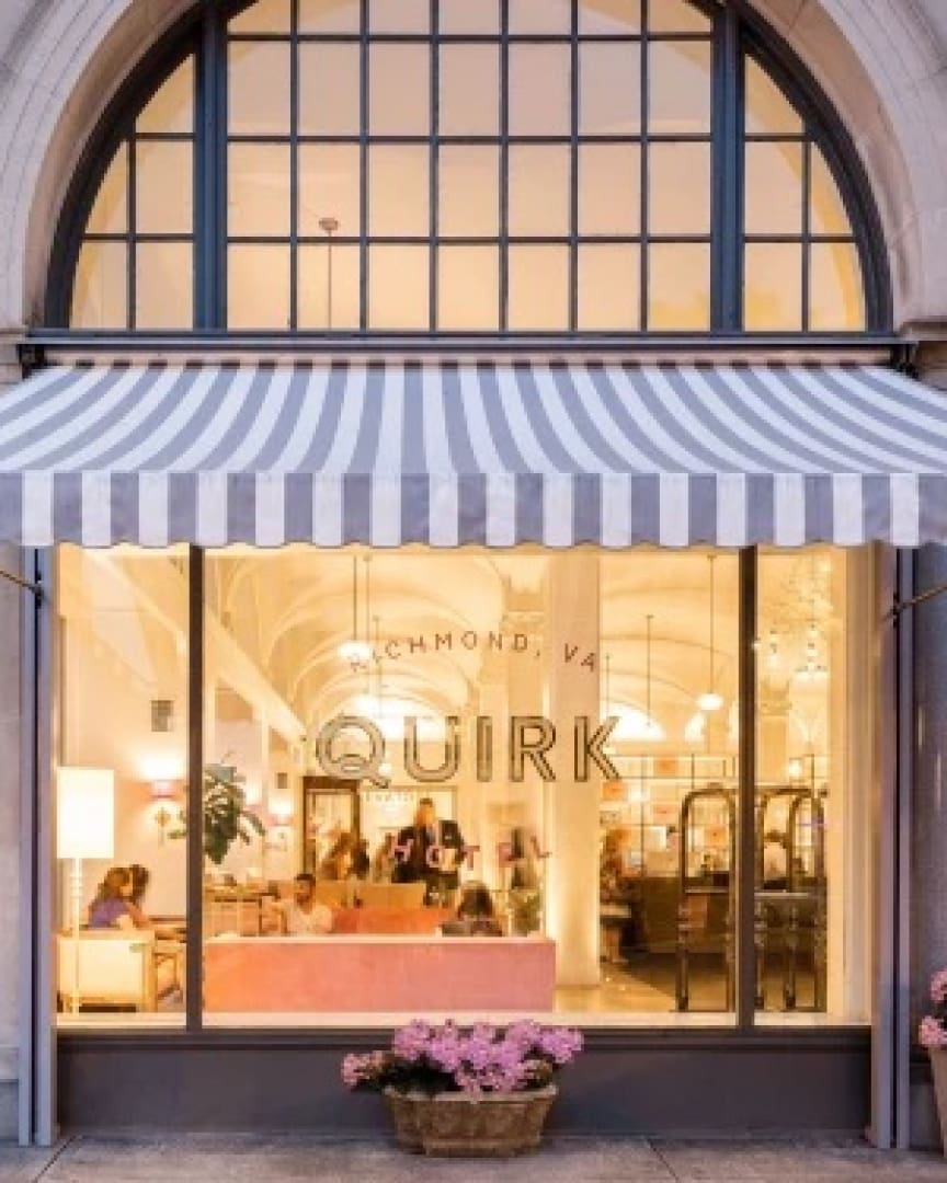 A large arched window with striped awning displays the warmly lit entrance of Quirk Hotel in Richmond, VA. Pink flowers are in a pot outside, and people are visible inside the stylish lobby.