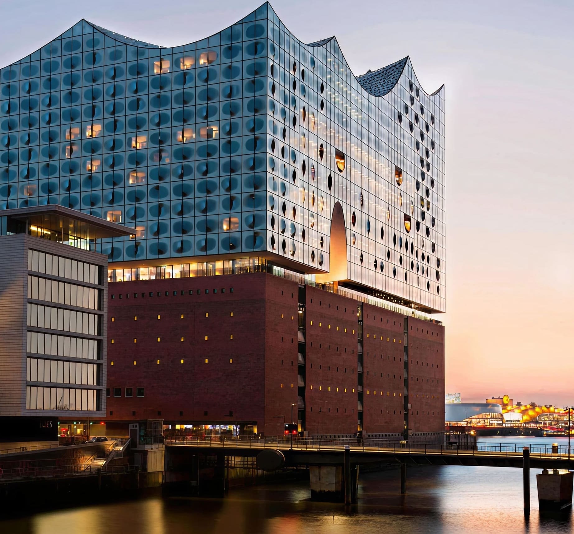 Modern glass concert hall with a wavy roof sits atop a historic brick warehouse by the water at sunset; some lights glow inside the building and the sky is clear.