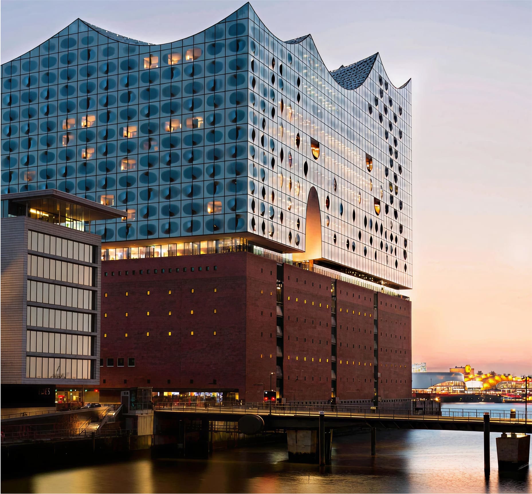 A modern concert hall with a glass, wave-shaped roof sits atop a brick warehouse by the water at sunset, reflecting light with its curved glass façade and circular window patterns.