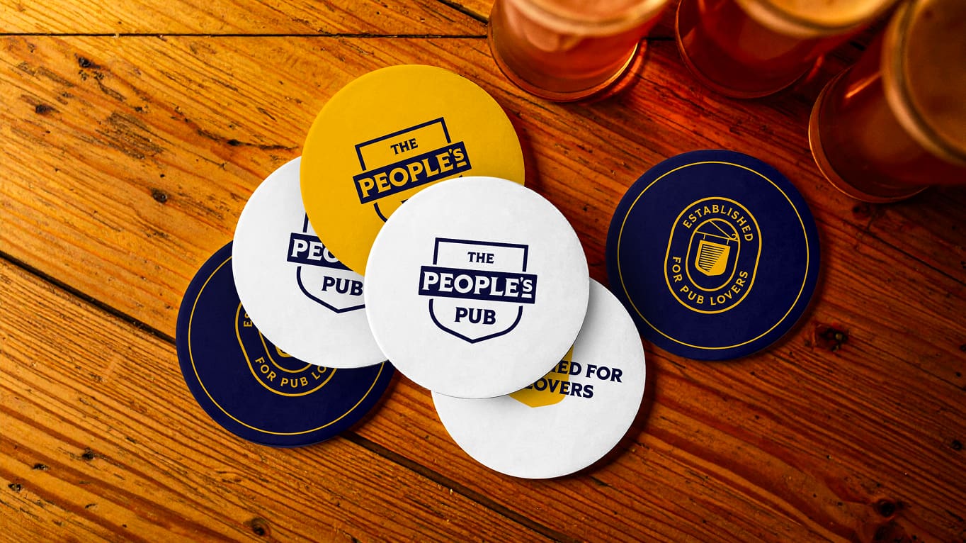 Round coasters with "THE PEOPLE'S PUB" and "ESTABLISHED FOR PUB LOVERS" logos are scattered on a wooden table near several glasses of amber-colored drinks.