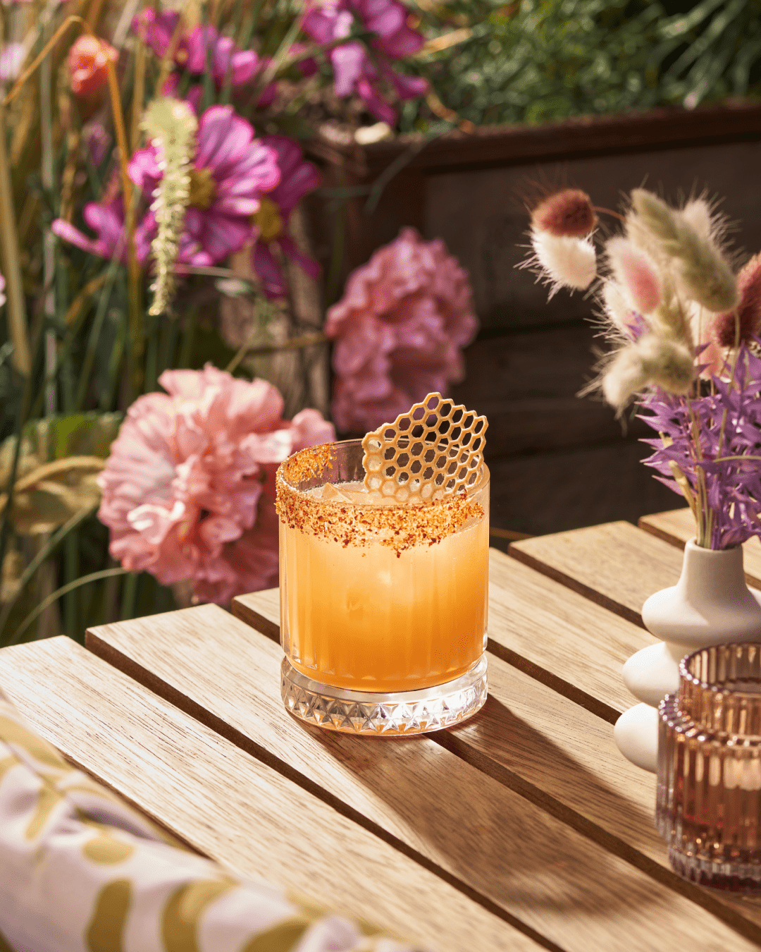 A cocktail with an orange hue and a chili-rimmed glass sits on a wooden table, garnished with a honeycomb-shaped decoration. Colorful flowers and greenery fill the background, creating a bright, summery outdoor scene.