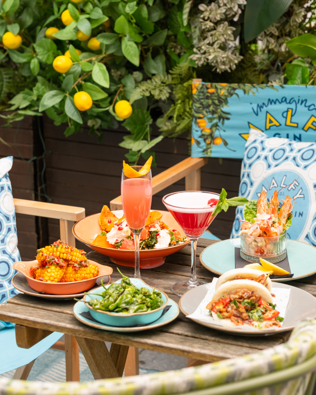 A colorful outdoor table spread with shrimp cocktail, grilled corn, stuffed buns, shishito peppers, salads, and two vibrant cocktails, set amid leafy plants and bright yellow lemons.