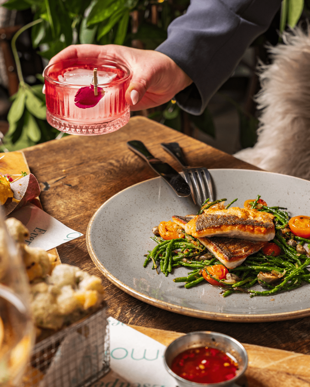 A hand holds a pink cocktail with a flower garnish above a plate of grilled fish, green vegetables, and cherry tomatoes on a rustic wooden table. Cutlery and additional dishes are visible nearby.