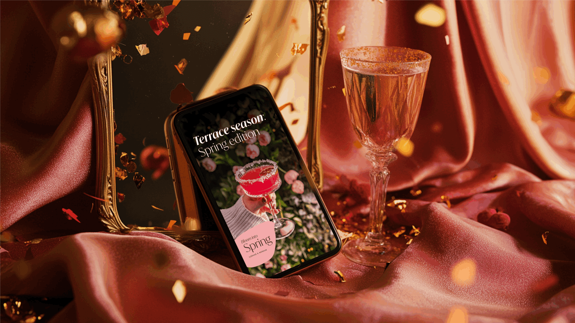 A smartphone displays a magazine cover featuring a pink cocktail. It’s placed on pink satin fabric next to a glass of pink drink, with gold confetti and a decorative mirror in the background.