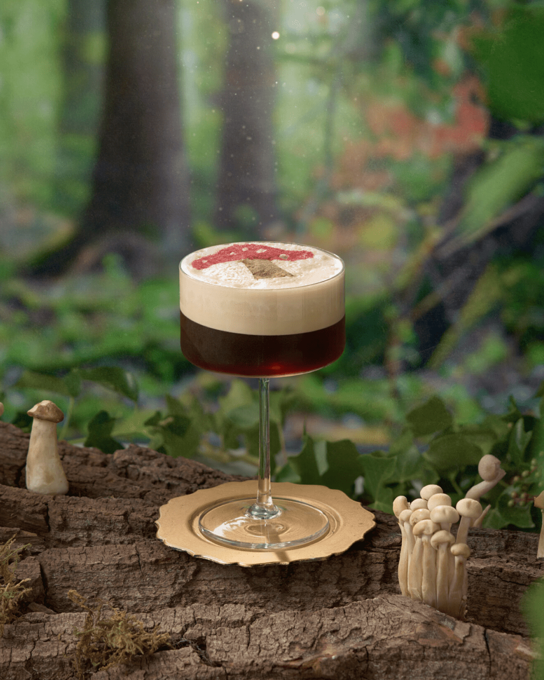A cocktail with a creamy foam top sits on a gold saucer atop a tree log, surrounded by small wild mushrooms, with a lush green forest in the blurred background.