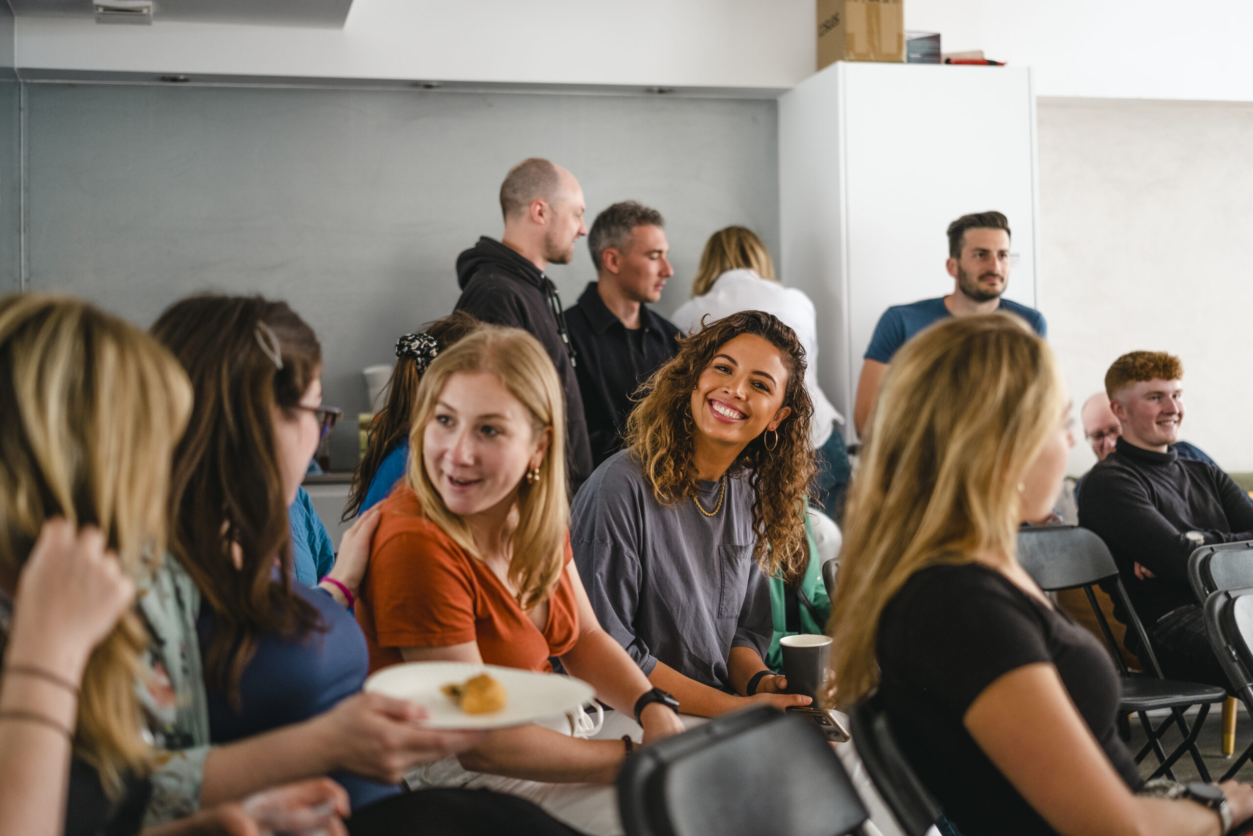 A group of people sit and chat in a classroom or meeting room. Some are smiling and holding drinks, while others stand in the background, engaged in conversation. The atmosphere appears friendly and relaxed.