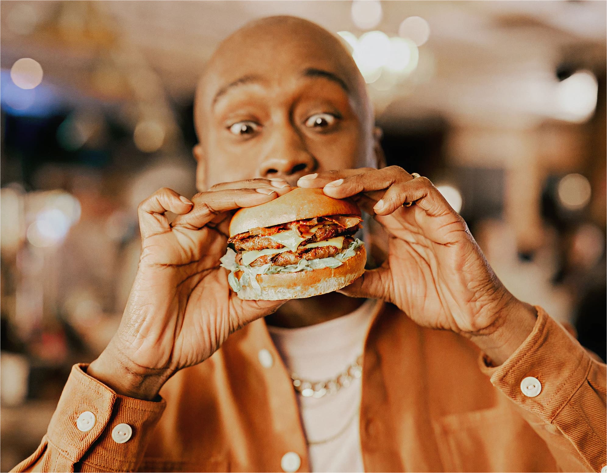 A person in a brown jacket holds a large hamburger with both hands, looking at it excitedly with wide eyes. The background is softly blurred, highlighting the focus on the person and the burger.