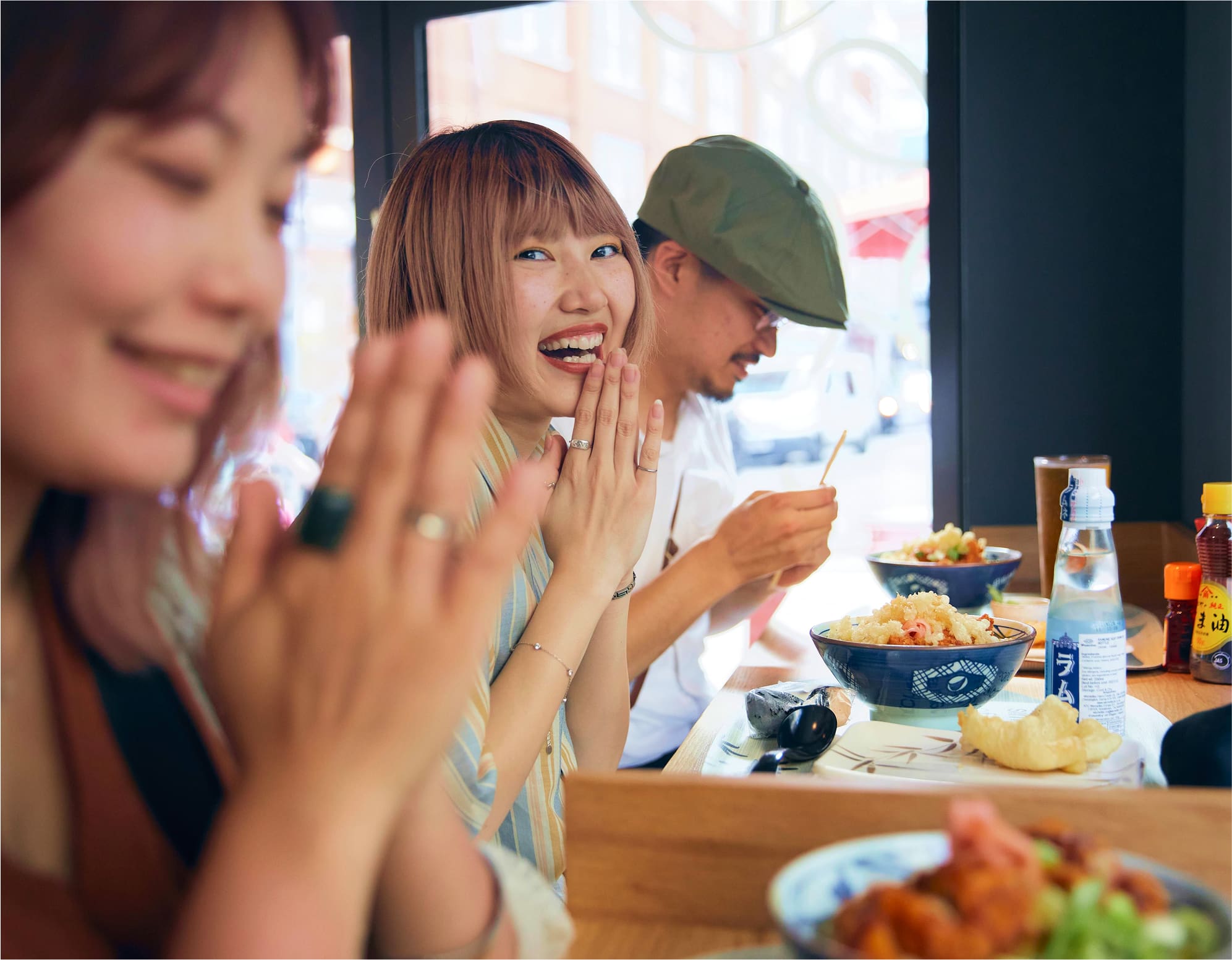 Three people sit at a restaurant table with bowls of food, smiling and clasping their hands in gratitude. The atmosphere is casual and warmly lit, with a city street visible through the window behind them.
