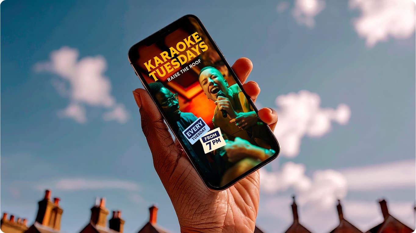 A hand holds up a smartphone displaying an event flyer for "Karaoke Tuesdays" with people singing, scheduled every Tuesday from 7 PM, against a bright sky with blurred rooftop silhouettes.