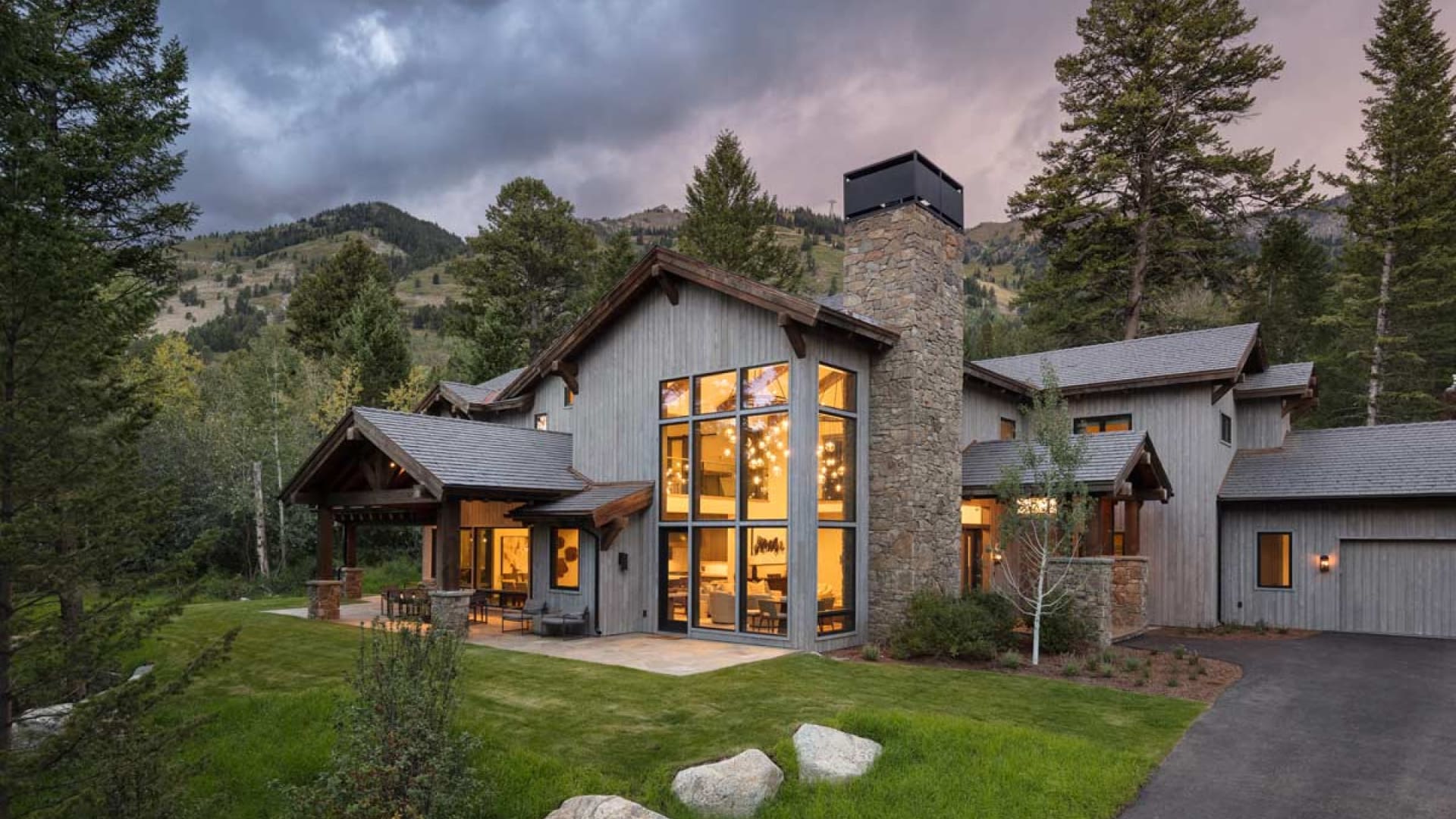 A modern rustic house with large windows and wood siding is surrounded by trees and mountains at dusk. Warm light glows from inside, highlighting the spacious interior. The home features a stone chimney and manicured lawn.