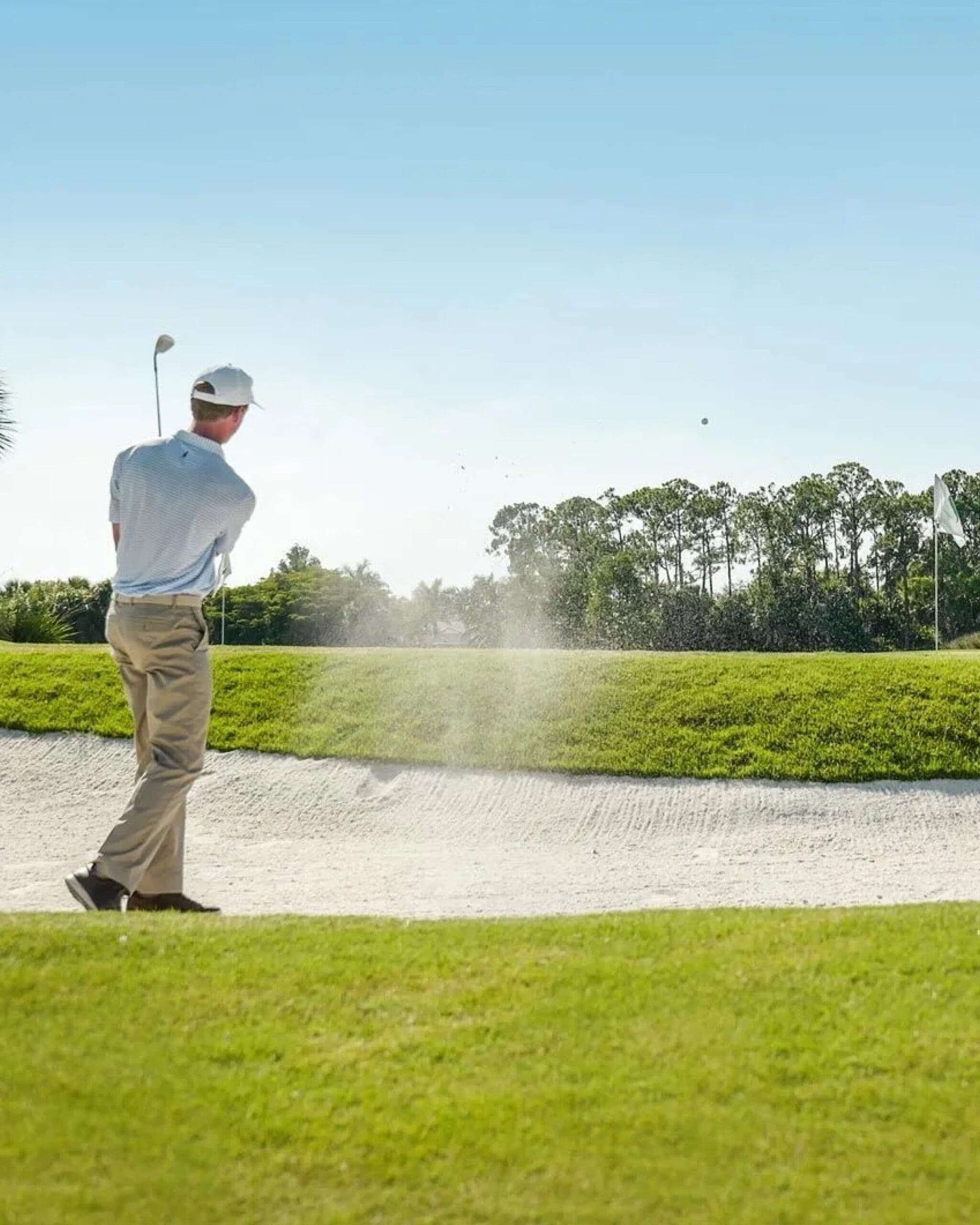 A golfer in a white cap and beige pants hits a shot from a sand bunker toward a green, with sand flying and a golf ball in midair under a clear blue sky.