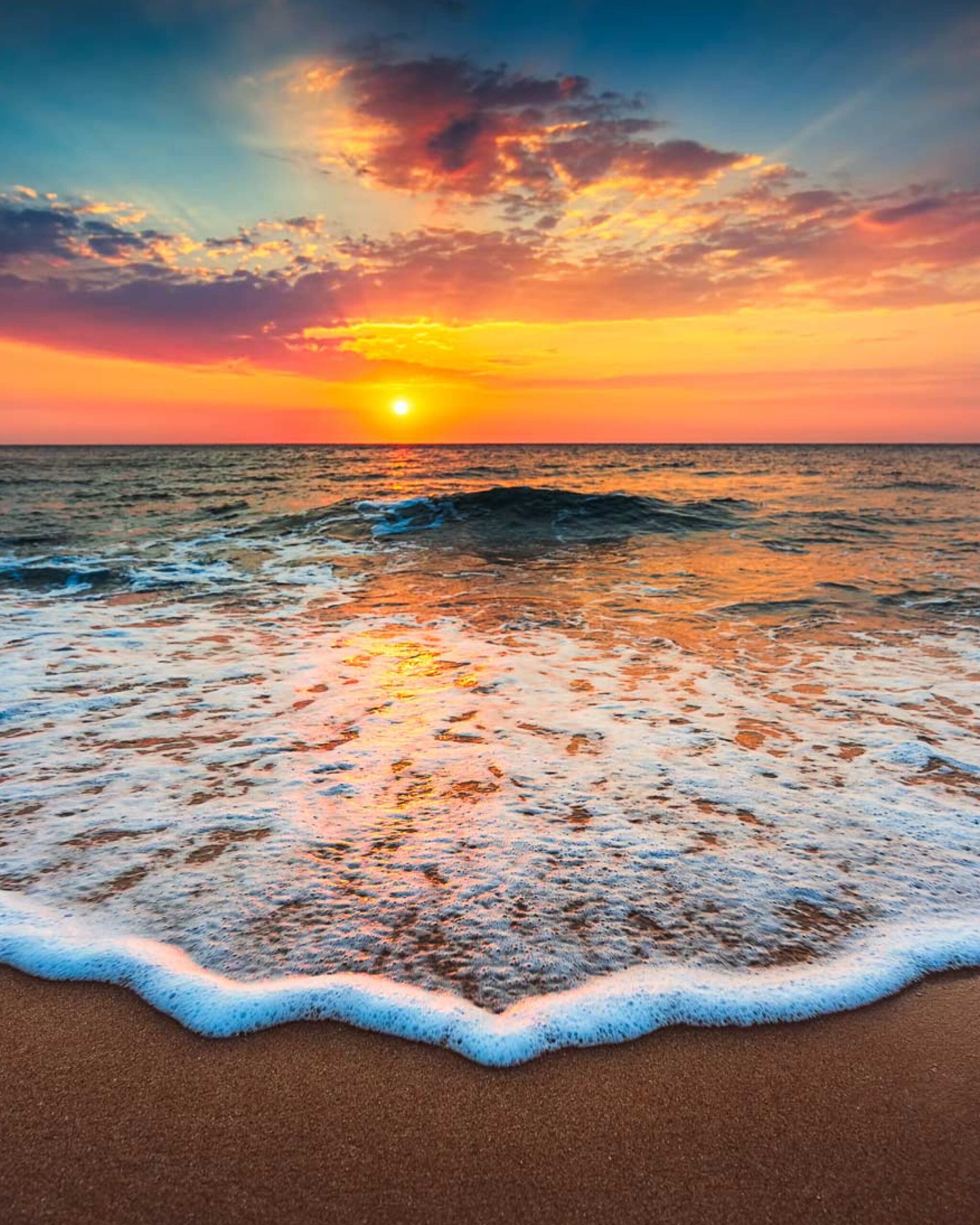 Sunset over a calm ocean, with colorful clouds in the sky and gentle waves washing onto a sandy beach in the foreground.
