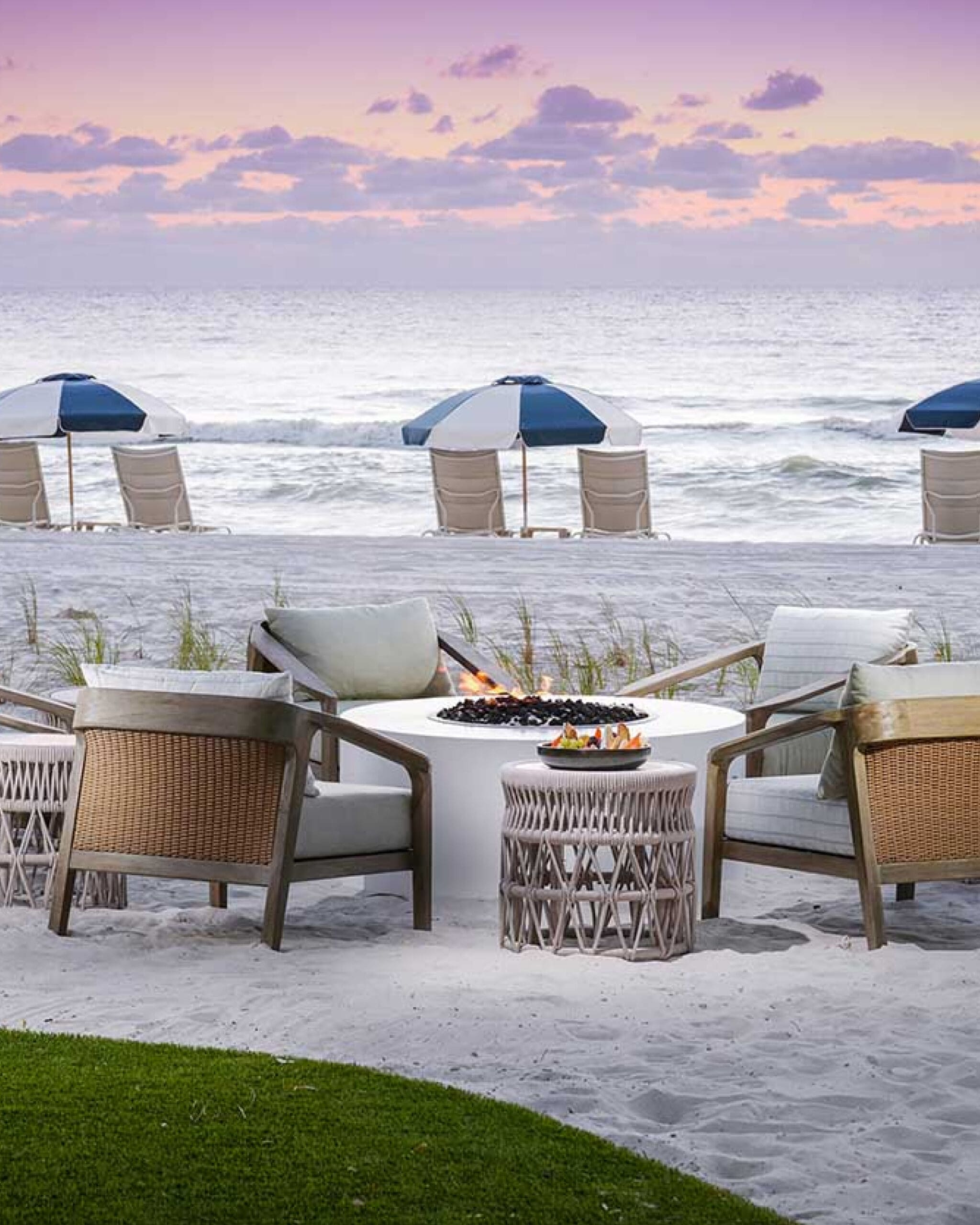 Four cushioned chairs surround a fire pit table on a sandy beach. In the background, lounge chairs and blue-and-white umbrellas face the ocean under a purple and orange sunset sky.