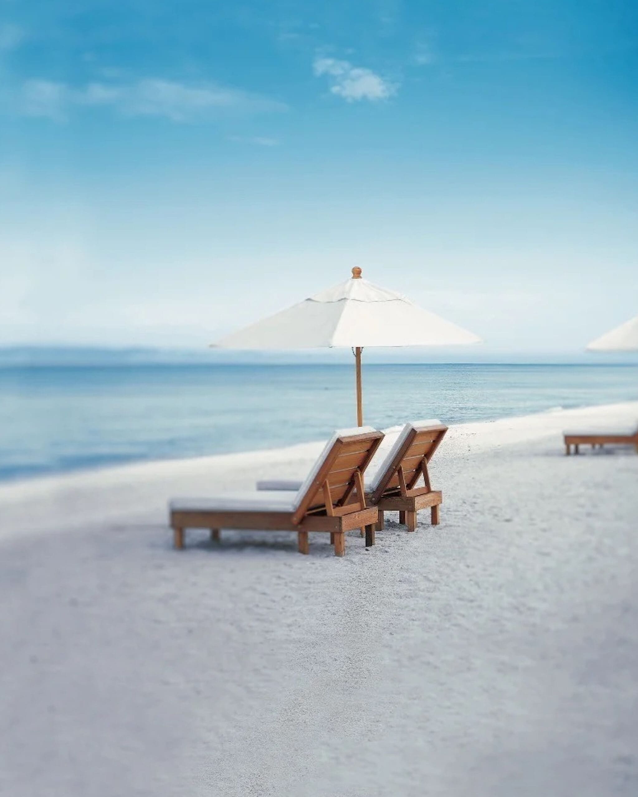 Two wooden lounge chairs with cushions sit under a white umbrella on a sandy beach, facing a calm blue ocean under a clear sky. The scene is peaceful and inviting.