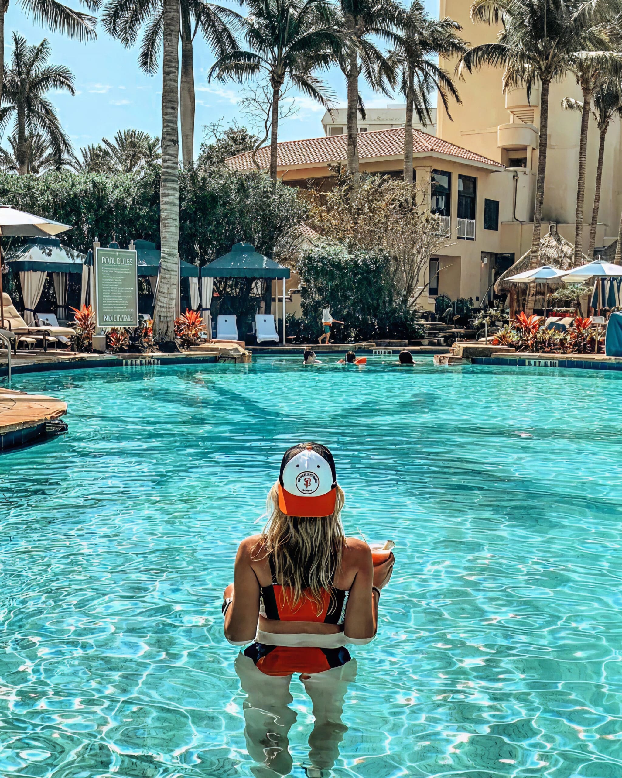A woman in a red bikini and a baseball cap stands waist-deep in a sunny hotel pool, facing away. Tropical plants, lounge chairs, and a yellow resort building with palm trees are visible in the background.