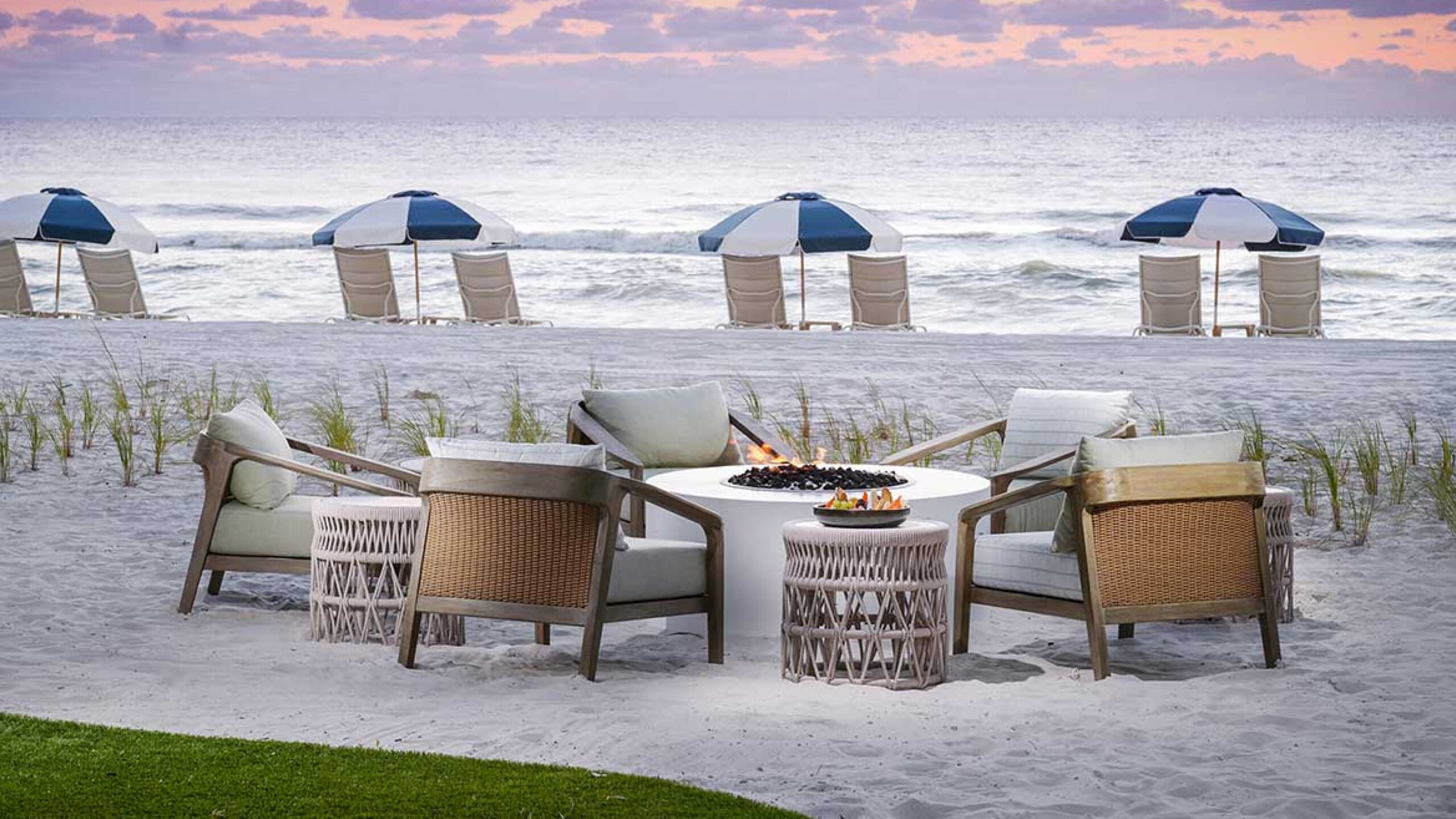A beachside seating area with five cushioned chairs around a round fire pit on white sand, overlooking the ocean. In the background, lounge chairs and blue-and-white umbrellas face the water at sunset.